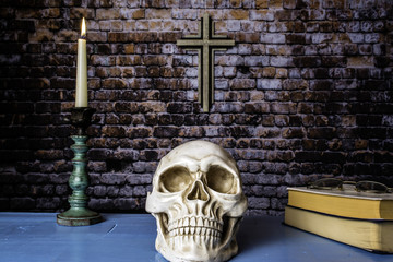 human skull bibles and lit candle in antique candle holder on blue table with wooden cross on brick wall background