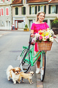 Beautiful blond woman wearing bright pink dress, riding a bicycle with big colorful bouquet of flowers in basket and holding leashes with two small dogs. Image taken in Lausanne, Switzerland