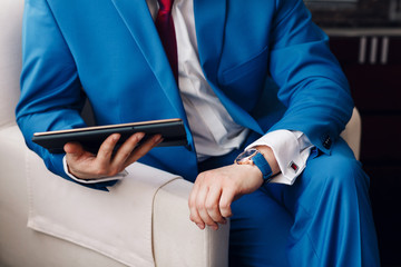 Businessman keep a digital tablet in hand whilst sitting on a sofa in a blue suit. on hand expensive mechanical watch with leather strap. shirt with cufflinks