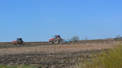 Fototapeta premium Two red tractors cultivate the field, agriculture, spring landscape, outskirts 