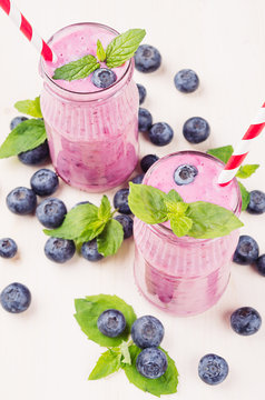 Freshly blended violet blueberry fruit smoothie in glass jars with straw, mint leaves, berries. White wooden board background.