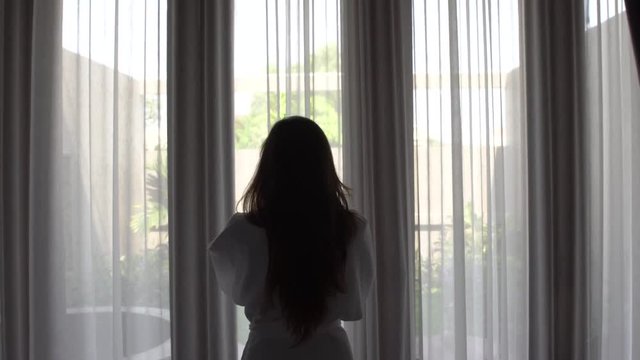 Young woman in bathrobe opens curtains and look at the garden standing near the window in her home