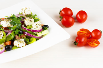 Greek salad with fresh tomato, cucumber, red onion, basil, feta cheese, black olives, Italian herbs and olive oil in white dish on white wooden table.