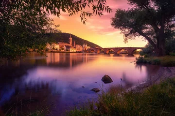 Heidelberg, Deutschland, verträumte Sonnenuntergangsfarben über dem Neckar mit der Altstadt und der Brücke, umrahmt von Baumsilhouetten © Smileus