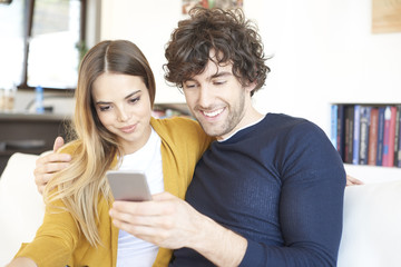 Getting some good news on mobile phone. Shot of a lovely young couple using smartphone while relaxing at home. 