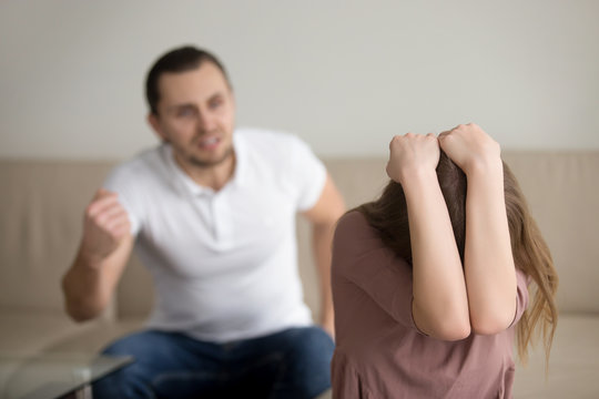 Aggressive mad husband threatening his wife with fist, angry boyfriend abusing girlfriend at home, lady is afraid and hiding head with hands, domestic violence awareness, physical abuse