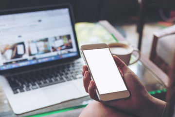 Mockup image of business woman holding mobile phone with blank white screen with laptop on wooden table in cafe