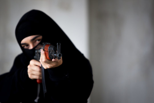 Unidentified man in black suit with mask is aiming assault rifle close up.