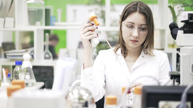 Front view of a woman scientist in a lab coat and protective glasses pouring liquid with a pipette from one test tube to another. Locked down real time medium shot
