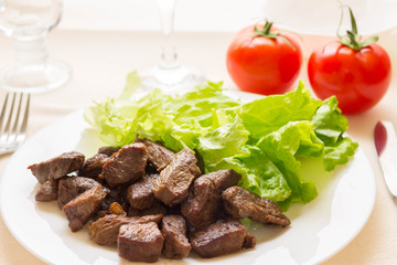 Fried beef goulash with lettuce leaves on a white plate and tomatoes in the background. Tasty and nutritious dish.