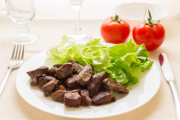 Fried beef goulash with lettuce leaves on a white plate and tomatoes in the background.