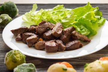 Fried beef goulash with lettuce leaves on a white plate and frozen vegetables. Tasty and nutritious dish.