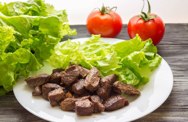 Fried beef goulash with lettuce leaves on a white plate and tomatoes in the background. Tasty and nutritious dish.
