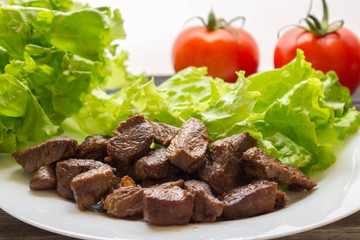 Fried beef goulash with lettuce leaves on a white plate and tomatoes in the background. Tasty and nutritious dish.