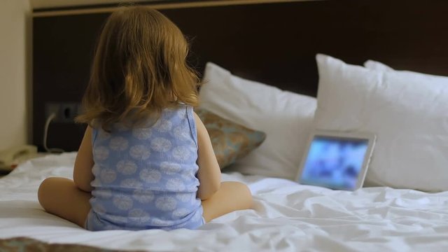 A child is sitting in the bedroom on the bed looking at the phone, tablet, computer.