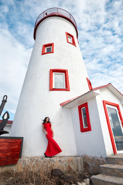 Sexy girl in red dress stands near the lighthouse on a Sunny summer day. Beautiful summer weather, vacation at sea. The woman on the background of lighthouse and blue sky with white clouds