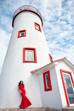 Sexy girl in red dress stands near the lighthouse on a Sunny summer day. Beautiful summer weather, vacation at sea. The woman on the background of lighthouse and blue sky with white clouds