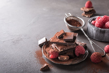 Broken chocolate pieces and raspberries with cocoa powder on metal plate on brown background with copy space. Dark photo.