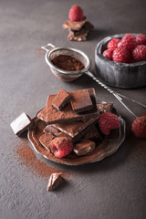Broken chocolate pieces and raspberries with cocoa powder on metal plate on brown background with copy space. Dark photo.