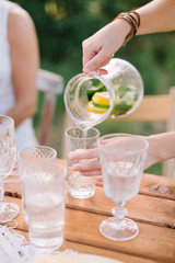 picnic, people, summer, holiday concept - female hands pouring lemonade from the decanter into a glass beaker, a jug with slices of lemon, lime, mint leaves, water, decorated table, selective focus