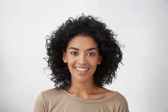 Close up shot of pretty girl with perfect teeth and dark clean skin having rest indoors, smiling happily after received good positive news. Beautiful young woman with Afro hairstyle relaxing at home