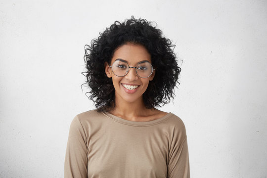 Carefree and relaxed pretty young mixed race female wearing big round eyeglasses smiling broadly, feeling excited about spending vacations abroad, standing isolated at white blank studio wall