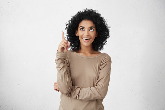 Look At That! Indoor Studio Portrait Of Cheerful Attractive Casually Dressed Young Female With Curly Hair Pointing Her Index Finger Up, Indication Something Interesting, Having Happy Excited Look