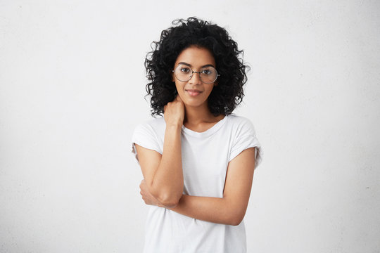 Cropped shot of beautiful friendly cute smiling young African American woman posing with closed posture, slightly smiling at camera, having shy look, wearing spectacles. Human emotions and gestures