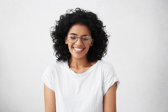 Portrait of charismatic and charming young African woman with curly hair wearing sylish spectacles, smiling widely, narrowing her eyes in expectation of surprise from her boyfriend, looking happy