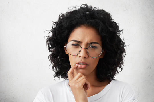Serious attractive young dark-skinned European woman wearing white casual t-shirt and round glasses keeping finger on her lips, having thoughtful indecisive look, thinking about something important