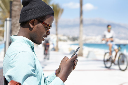 Profile picture of attractive young African American man in hat and shades playing video games using online app on smart phone, enjoying free wireless high-speed internet connection on urban beach