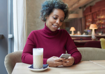 Indoor portrait of good-looking casually dressed African American lady sitting in coffee house during lunchtime on her own, enjoying friendly online communication via smartphone with happy look.