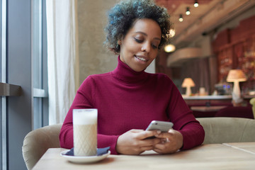 Closeup of beautiful dark skinned woman with colored curly hair spending time alone in cafe holding...