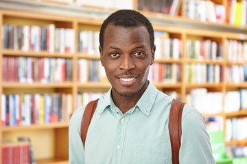 People, knowledge and education concept. Attractive young Afro American male with knapsack came to bookstore to buy science fiction or professional literature, standing against shelves background