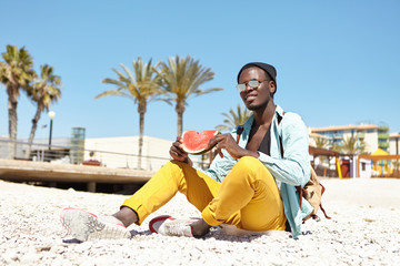 Outdoor portrait of handsome young African American man with knapsack sitting on pebble beach wearing comfortable stylish clothing, backpack and shades, holding slice of watermelon in his hands