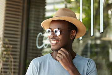 Happy dark-skinned European male youngster in hat and sunglasses relaxing at sidewalk cafe on sunny summer day, waiting for his lunch, smiling cheerfully, enjoying nice weather and leisure time