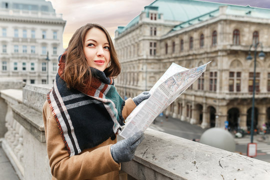 A stylish woman in a coat with a scarf is guided by a map in the center of a European city, the concept of travel
