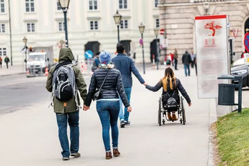 Foto op Plexiglas A man with a woman goes by the hand through the streets of Vienna against the background of another couple in love in a wheelchair, disabled love concept © EdNurg