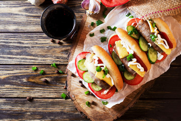 Snack, fast food, hot dogs with Bavarian hunting sausages, tomato, cucumber, green onion, cheese, ketchup, mayonnaise and a glass of cola or dark beer on a wooden  rustic background