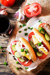 Snack, fast food, hot dogs with Bavarian hunting sausages, tomato, cucumber, green onion, cheese, ketchup, mayonnaise and a glass of cola or dark beer on a wooden  rustic background