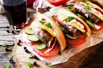 Snack, fast food, hot dogs with Bavarian hunting sausages, tomato, cucumber, green onion, cheese, ketchup, mayonnaise and a glass of cola or dark beer on a wooden  rustic background