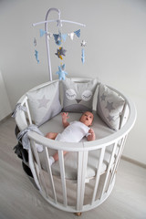 newborn lies in the round white bed with mobile and toys