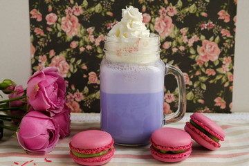 Purple coffee in stylized mason jar cup with macarons and roses and colorful decoration on background of floral pattern. Blueberry milk shake. Unicorn coffee. Unicorn food