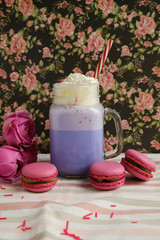 Purple coffee in stylized mason jar cup with macarons and roses and colorful decoration on background of floral pattern. Blueberry milk shake. Unicorn coffee. Unicorn food