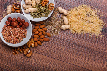 Assorted nuts in white bowl, plate on wooden surface. Top view with copy space