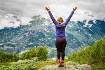 Rear view of young successful woman hiker open arms on mountain peak. Day cloudy scene, snow caped norwegian mountains and green trees canyon ahead person. Location: Odda, Rogaland, Norway, Europe.