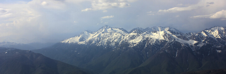 View from top of mountain peak. Green mountain valley in blue haze and snowy mountain range. Ravine and cloudy sky. Sochi, Russia.
