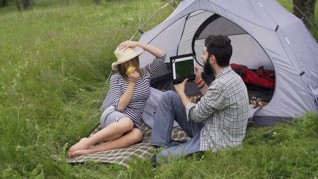  Happy couple enjoying the nature at camping trip using a tablet for selfie. Beautiful blonde girl eating an apple and posing for a photo at her boyfriend who filmed with a digital tablet.