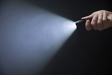 Black flashlight in the man's hand emerging from the right corner of the frame with a wide light beam, isolated on black background - obrazy, fototapety, plakaty