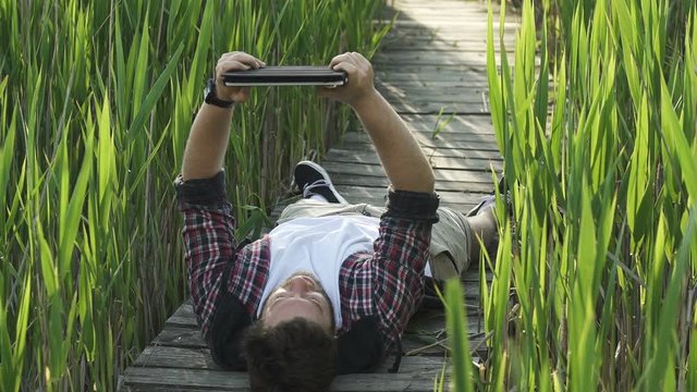  A young hipster man surfing Internet with a digital tablet computer and lying on the footpath of bulrush